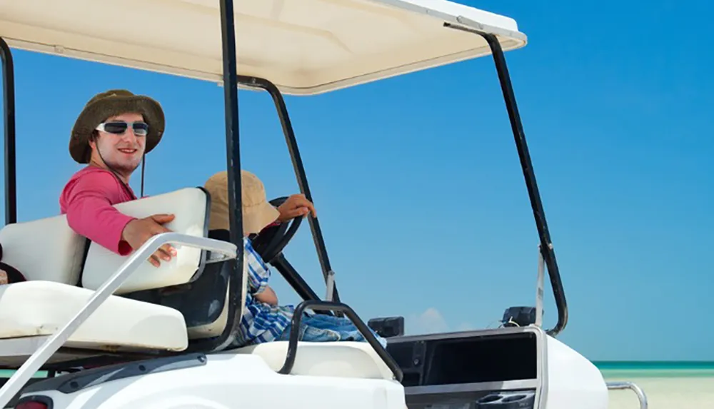 Two individuals are riding in a golf cart along a beautiful beach under a clear blue sky