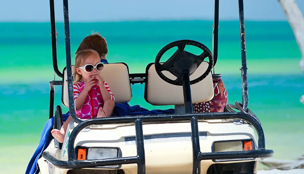 A child with sunglasses is sitting in the drivers seat of a stationary golf cart on a sunny beachfront