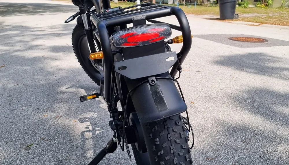 The image shows the rear view of a black electric bicycle with a red tail light and turn signals parked on a pavement