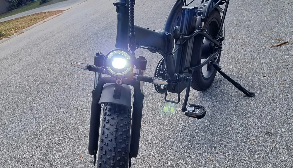 The image shows the front part of an electric bicycle with its headlight on parked on an asphalt road