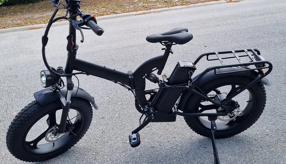 The image shows a black electric fat-tire bicycle parked on a road featuring a headlight a rear cargo rack and a mid-frame battery pack