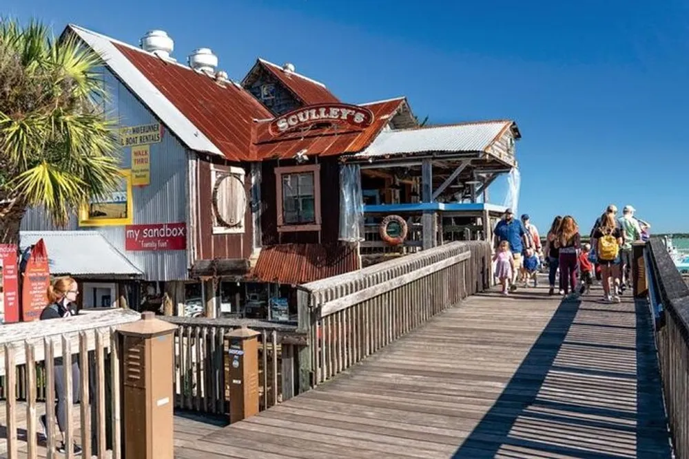 A rustic waterfront restaurant named Sculleys sits alongside a wooden boardwalk bustling with people under a clear blue sky