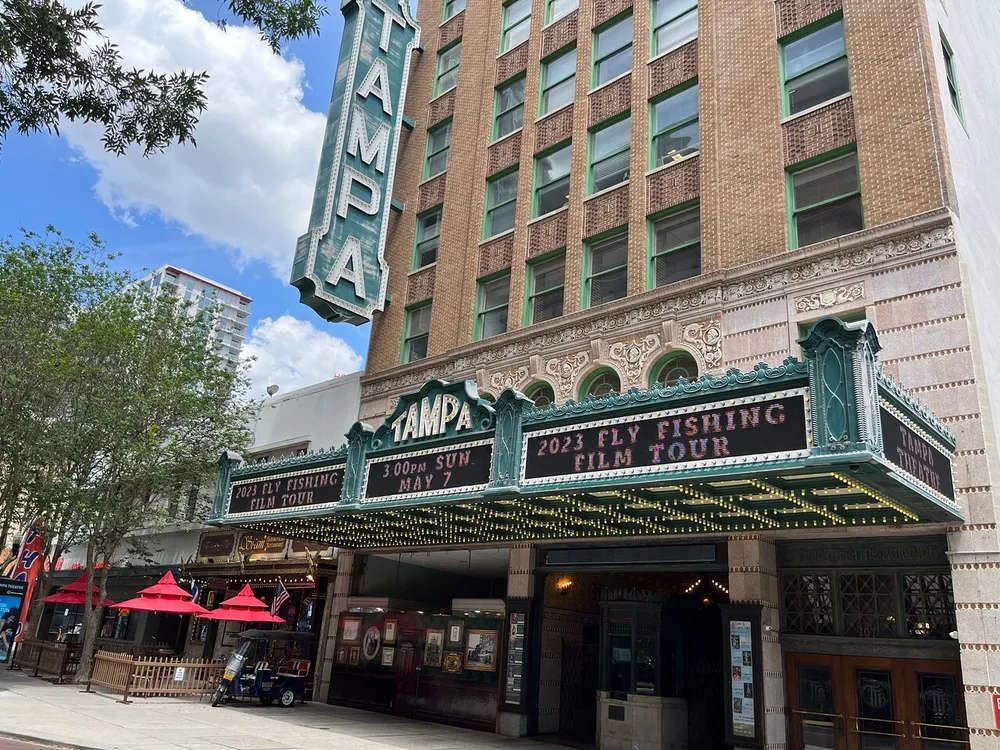 The image shows the historic Tampa Theatre with a marquee advertising a 2023 Fly Fishing Film Tour situated in a downtown area on a sunny day
