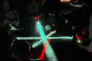 A group of people in a dimly lit environment are creating patterns on the floor with colorful lights, possibly from light sabers or similar illuminated devices.