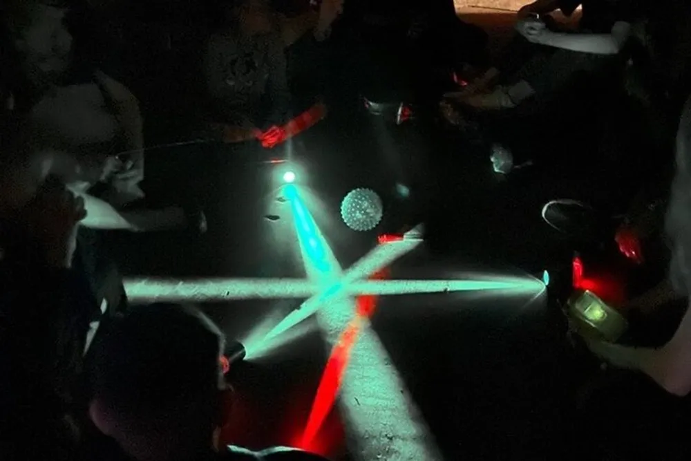 A group of people in a dimly lit environment are creating patterns on the floor with colorful lights possibly from light sabers or similar illuminated devices