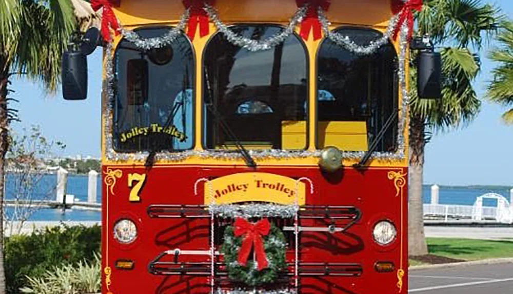 The image shows a red and yellow trolley bus adorned with festive holiday decorations including tinsel and a wreath parked in a sunny location with a view of the water and palm trees in the background