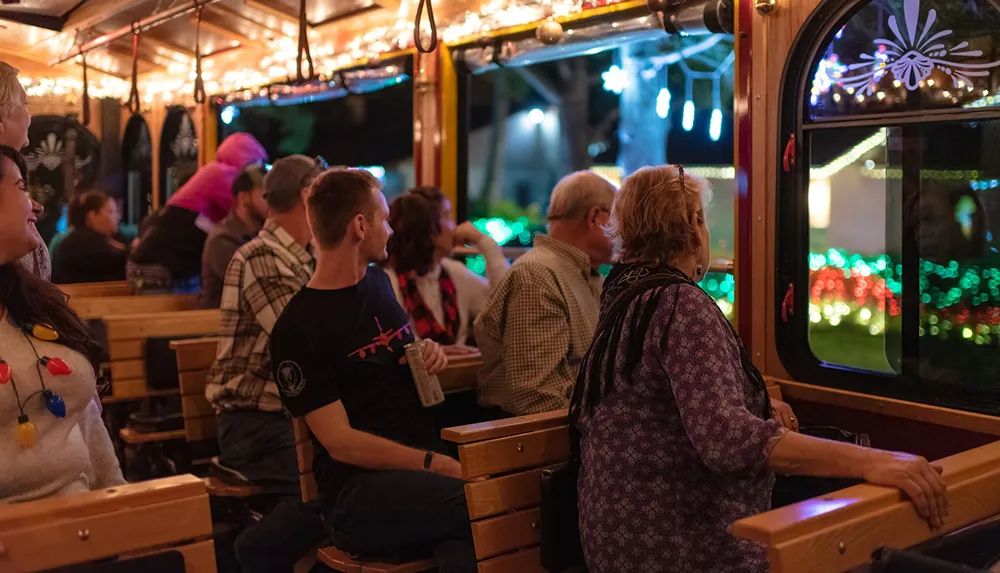 Passengers are enjoying a festive ride in a trolley adorned with twinkling lights looking out at colorful illuminations outside