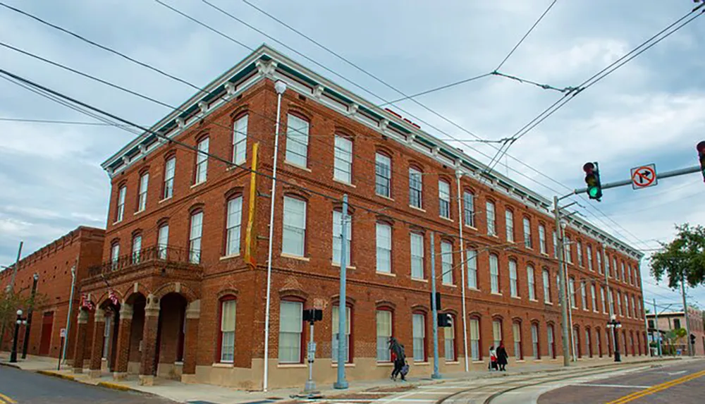 This image shows a three-story red-brick corner building with arched windows on the ground floor located in an urban area with overhead lines likely for electrical supply or transit as people walk by on the sidewalks