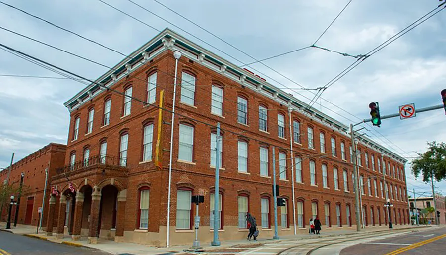 This image shows a three-story, red-brick corner building with arched windows on the ground floor, located in an urban area with overhead lines, likely for electrical supply or transit, as people walk by on the sidewalks.