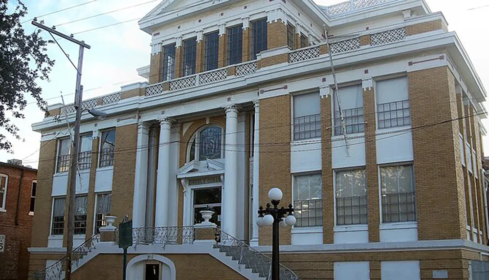 The image showcases a yellow brick building with a classical revival architectural style featuring large white columns and stairs leading up to a grand entrance