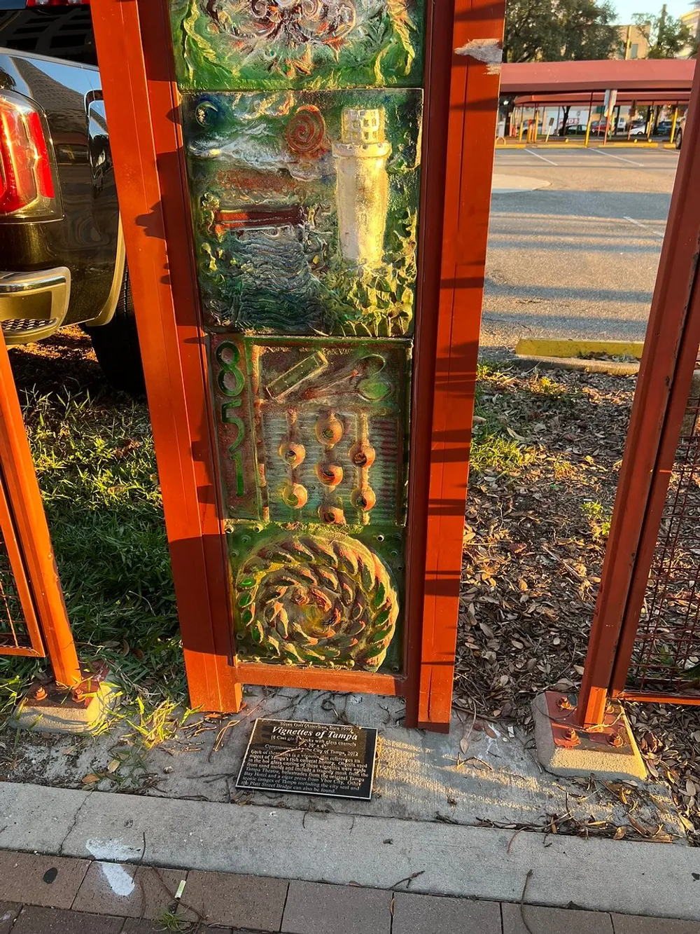 The image shows an ornate colorful sculptural panel within an orange frame installed outdoors near a sidewalk with a plaque titled Figurines of Tampa at its base