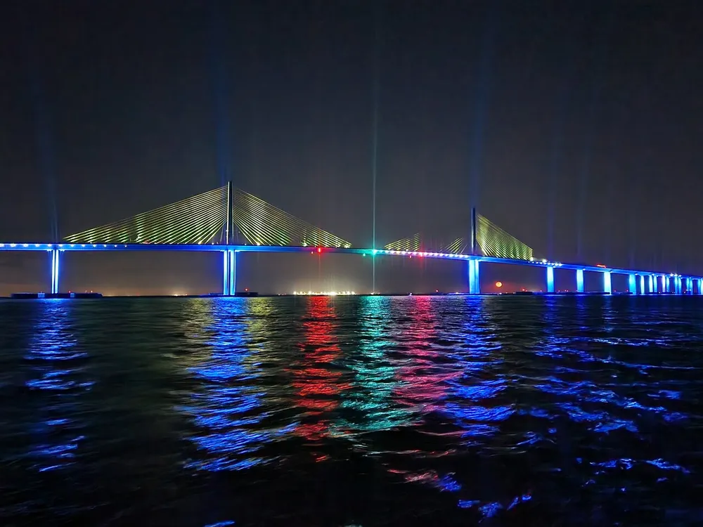 A long suspension bridge is illuminated with colorful lights at night reflecting on the waters surface
