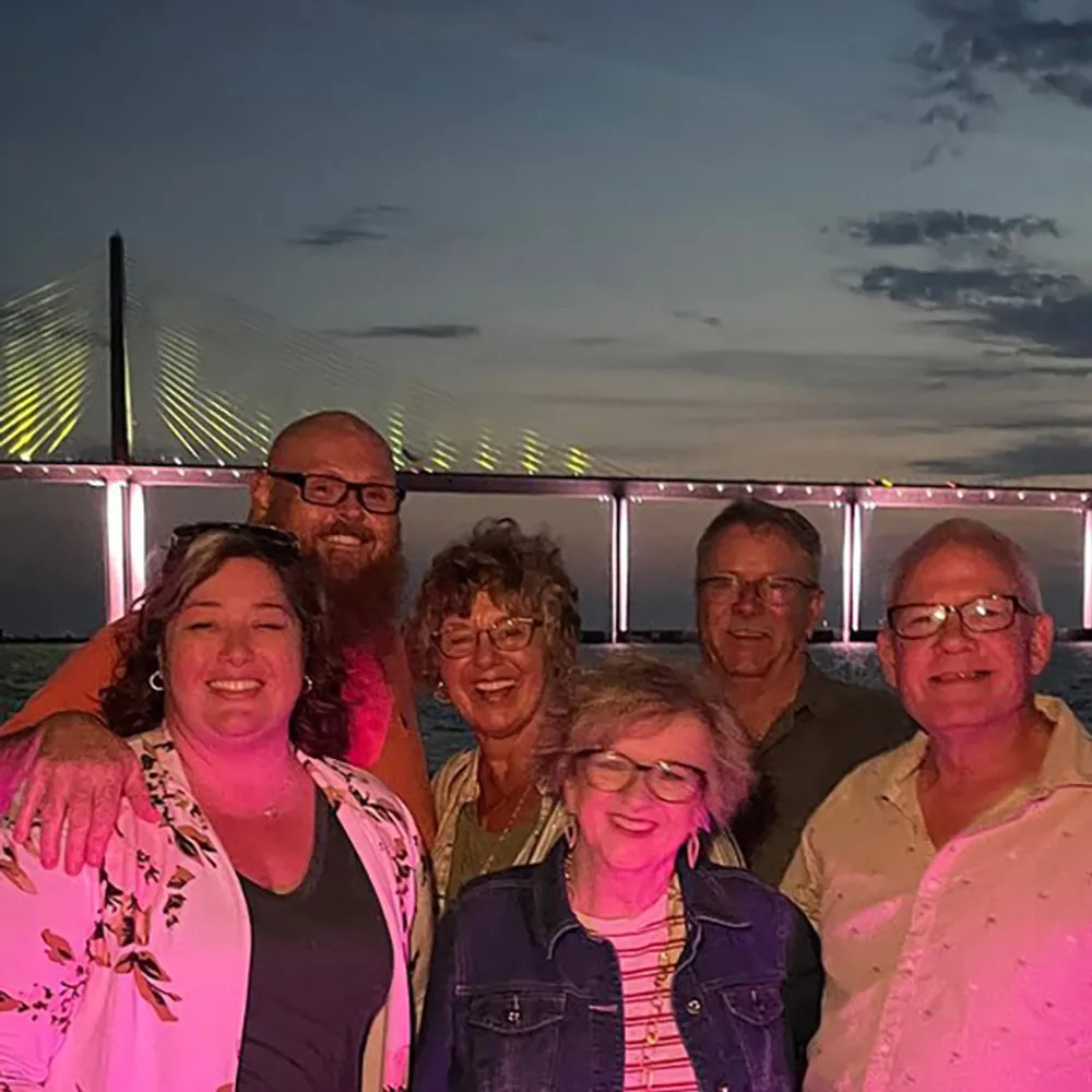 A group of six people is smiling in front of a lit-up bridge during dusk