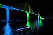 A brightly illuminated bridge casts vibrant green and blue lights onto the water at night, with a small boat passing underneath.