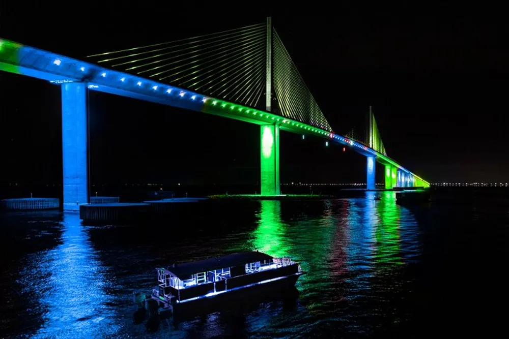 A brightly illuminated bridge casts vibrant green and blue lights onto the water at night with a small boat passing underneath