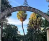 The image shows the rusted iron gate entrance with the sign OAKLAWN CEMETERY framed by tropical trees under a blue sky