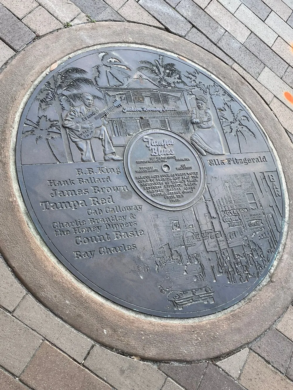 This image features a commemorative plaque embedded in the ground celebrating artists of the Tampa Blues genre with intricate engravings and names including BB King James Brown Ella Fitzgerald and Ray Charles
