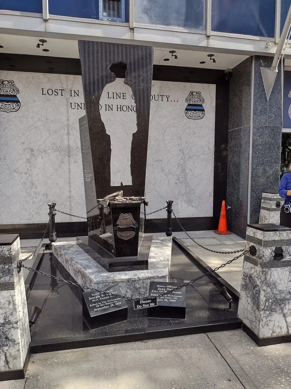This image shows a solemn memorial for fallen police officers featuring a black stone monument with an engraved silhouette inscriptions and adorned with commemorative plaques and a bronze hat and handcuffs set against a marbled backdrop with the inscription LOST IN LINE OF DUTY UNITED IN HONOR