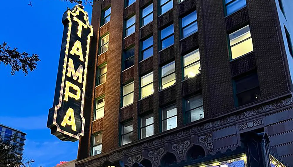 An illuminated vertical TAMPA sign adorns the side of a multi-story brick building in an urban setting during dusk
