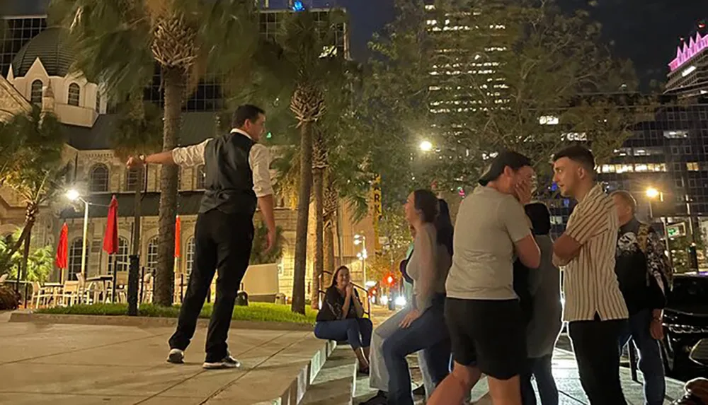A man in a vest and white shirt is performing or demonstrating something at night to an attentive group of onlookers in an urban palm-lined setting