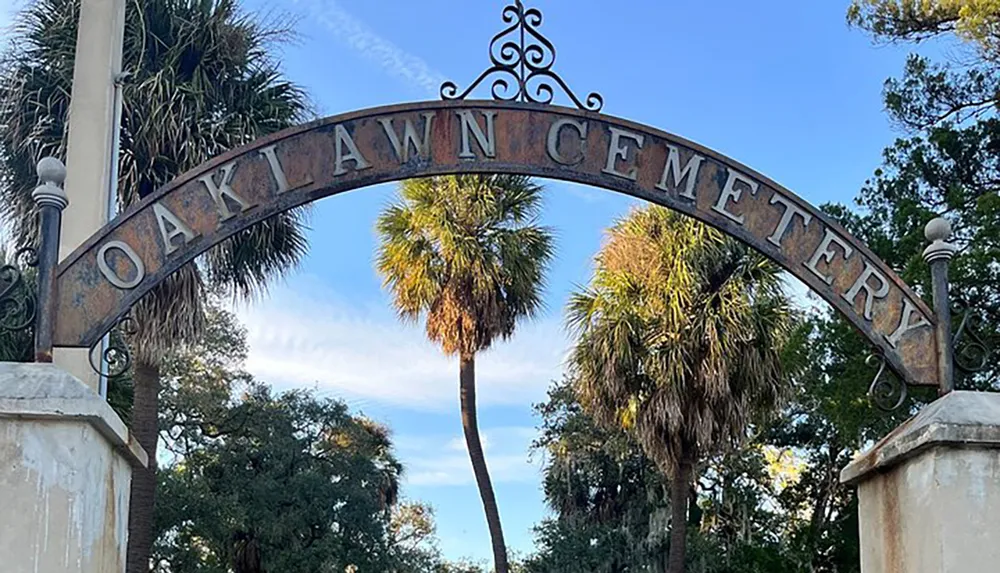 The image shows an arched metal sign with the inscription OAKLAWN CEMETERY framed by a blue sky and flanked by palm trees