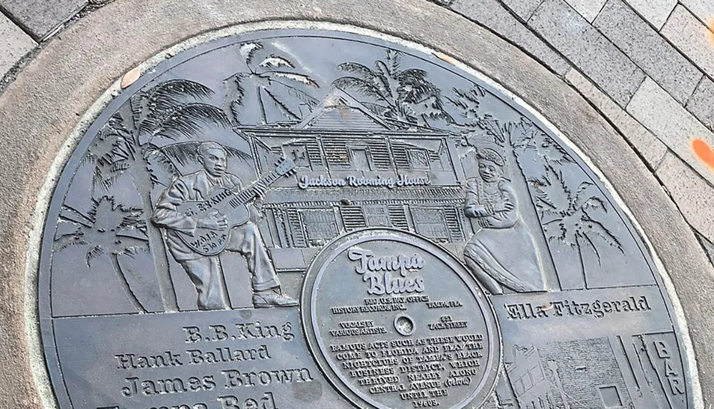 This image features a commemorative plaque embedded in the ground celebrating Tampa Blues with relief sculptures of musicians BB King and Ella Fitzgerald as well as the image of the Jackson Rooming House and text indicating historical significance