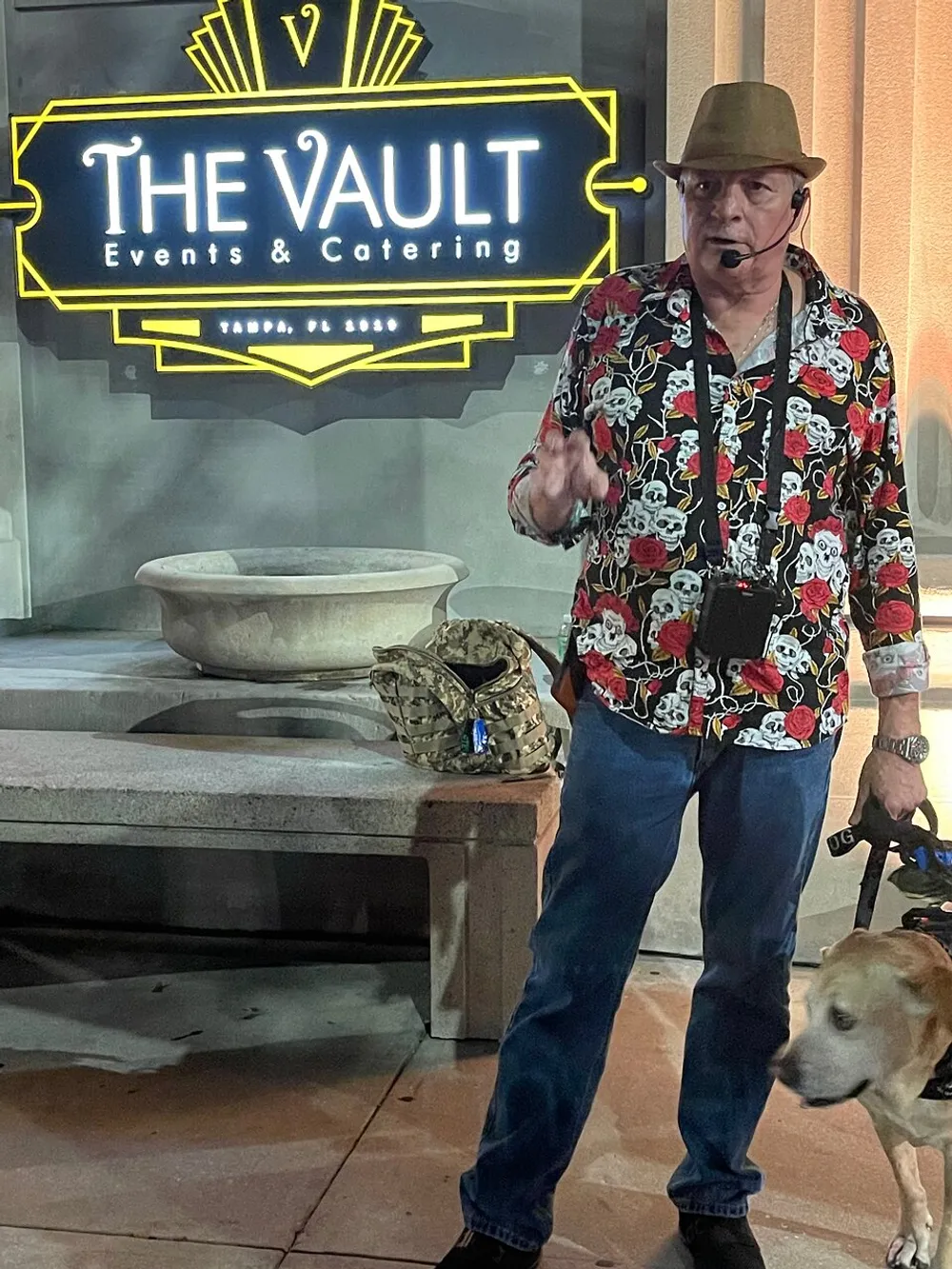 A person wearing a floral shirt and a hat is standing in front of a sign that reads THE VAULT Events  Catering with a dog walking by and a snake on a ledge to the left