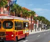 A trolley styled bus adorned with advertisements is parked on a sunny street lined with palm trees