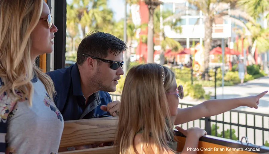 A family appears engaged and curious as a child points to something of interest outside of a trolley or bus they are seated in, under a bright and sunny sky.