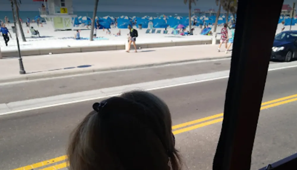 The image shows a person looking out towards a sunny beachside street lined with umbrellas as viewed from behind and somewhat obscured by a shadowed interior