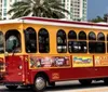 A trolley styled bus adorned with advertisements is parked on a sunny street lined with palm trees