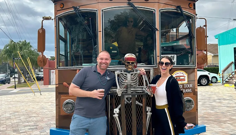 Two people are smiling and posing next to a skeleton figure in front of a trolley-style vehicle that advertises a Mystery Tour Crawl