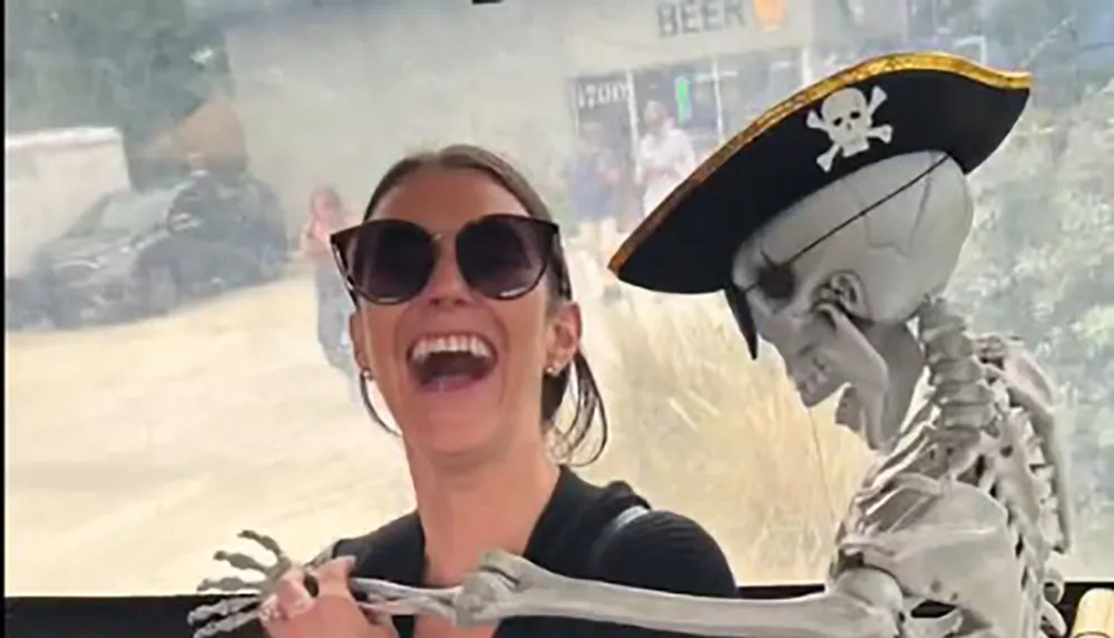 A joyful woman is wearing sunglasses and laughing while a skeleton wearing a pirate hat appears to put its arm around her