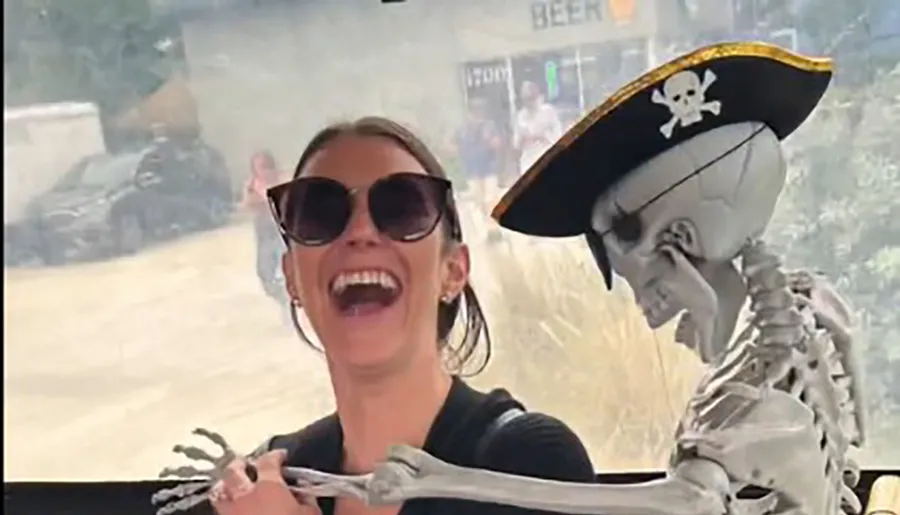 A joyful woman is wearing sunglasses and laughing while a skeleton wearing a pirate hat appears to put its arm around her.