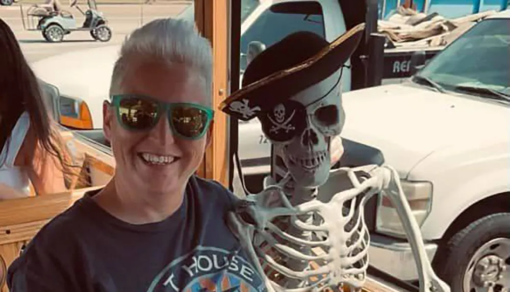 A person is smiling next to a skeleton wearing a pirate hat both of them sporting sunglasses in a setting with vehicles in the background