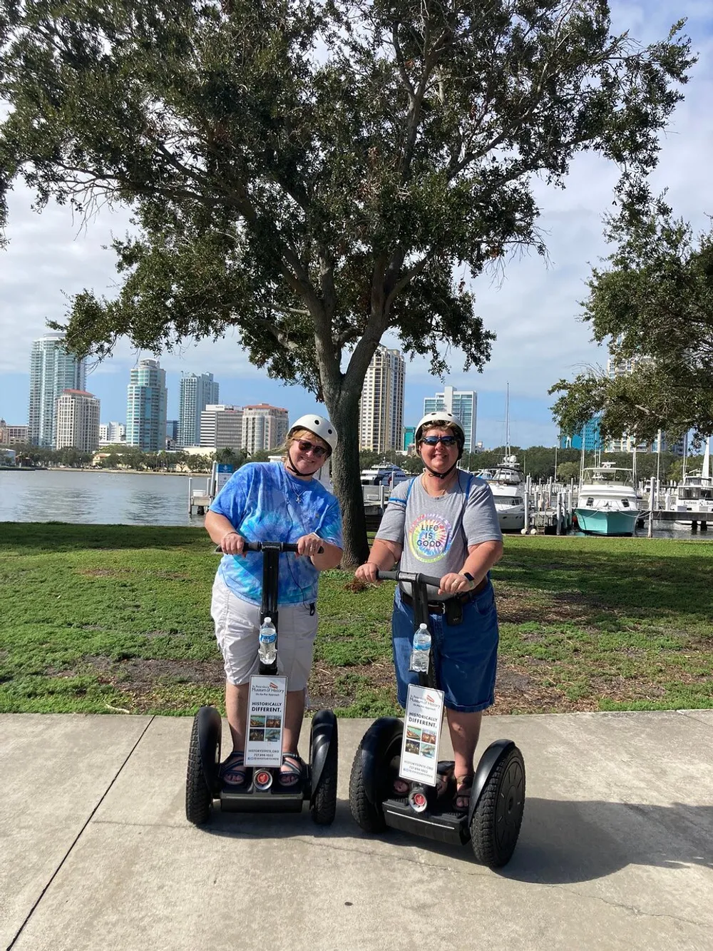 Two people are standing on Segways enjoying a sunny day outdoors with a backdrop of skyscrapers and a marina