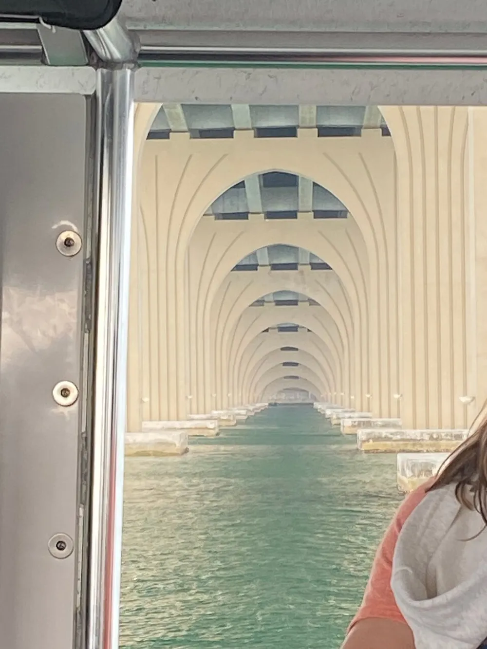 The image shows the underbelly of a bridge taken from a boat revealing a vanishing point perspective of repetitive structural arches over water with part of a person visible on the right