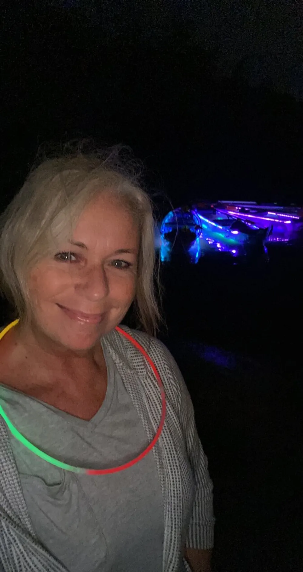 A smiling person is taking a selfie at night with colorful glowing lights in the background wearing a glow necklace