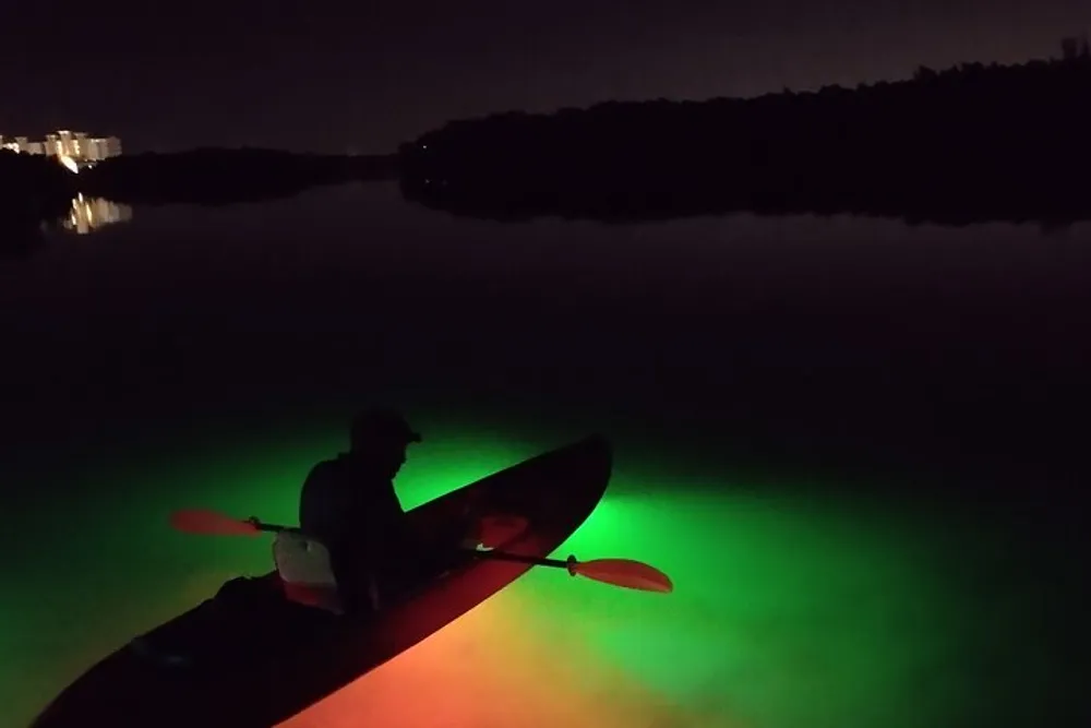 A person is kayaking at night on calm water illuminated by a bright green underwater light