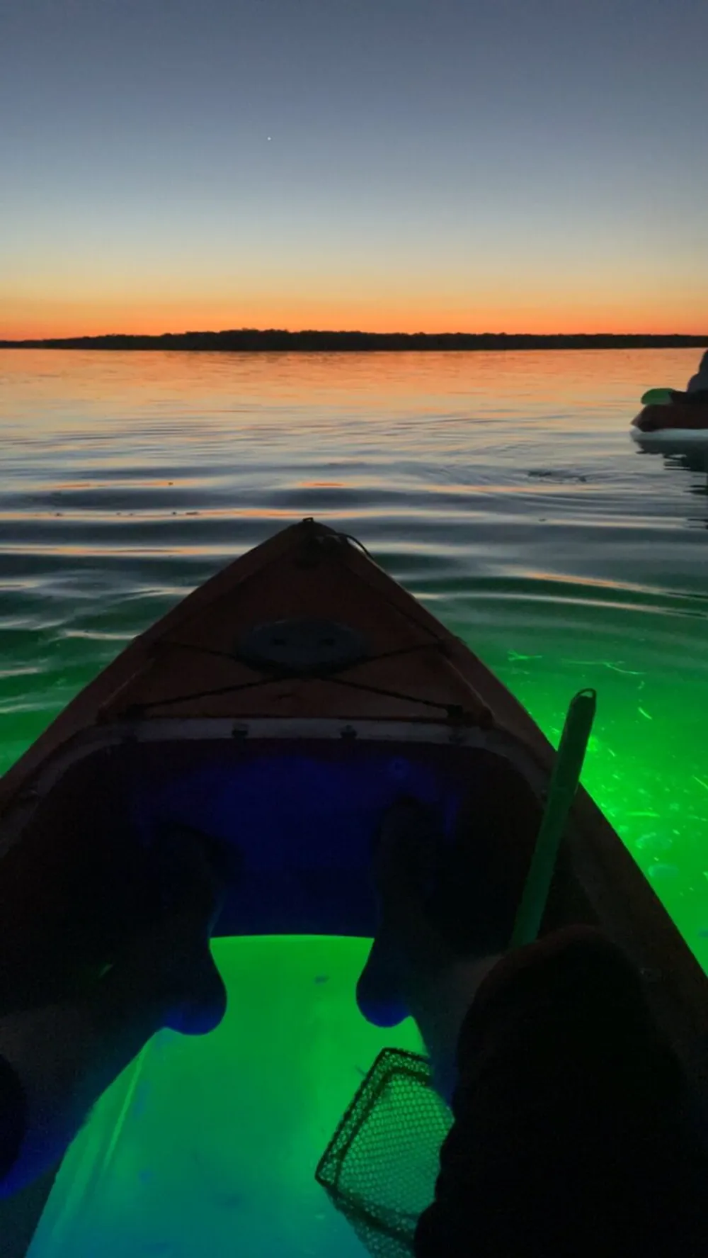 A person is kayaking on calm waters at dusk with a vivid display of sunset colors above and illuminated green water below