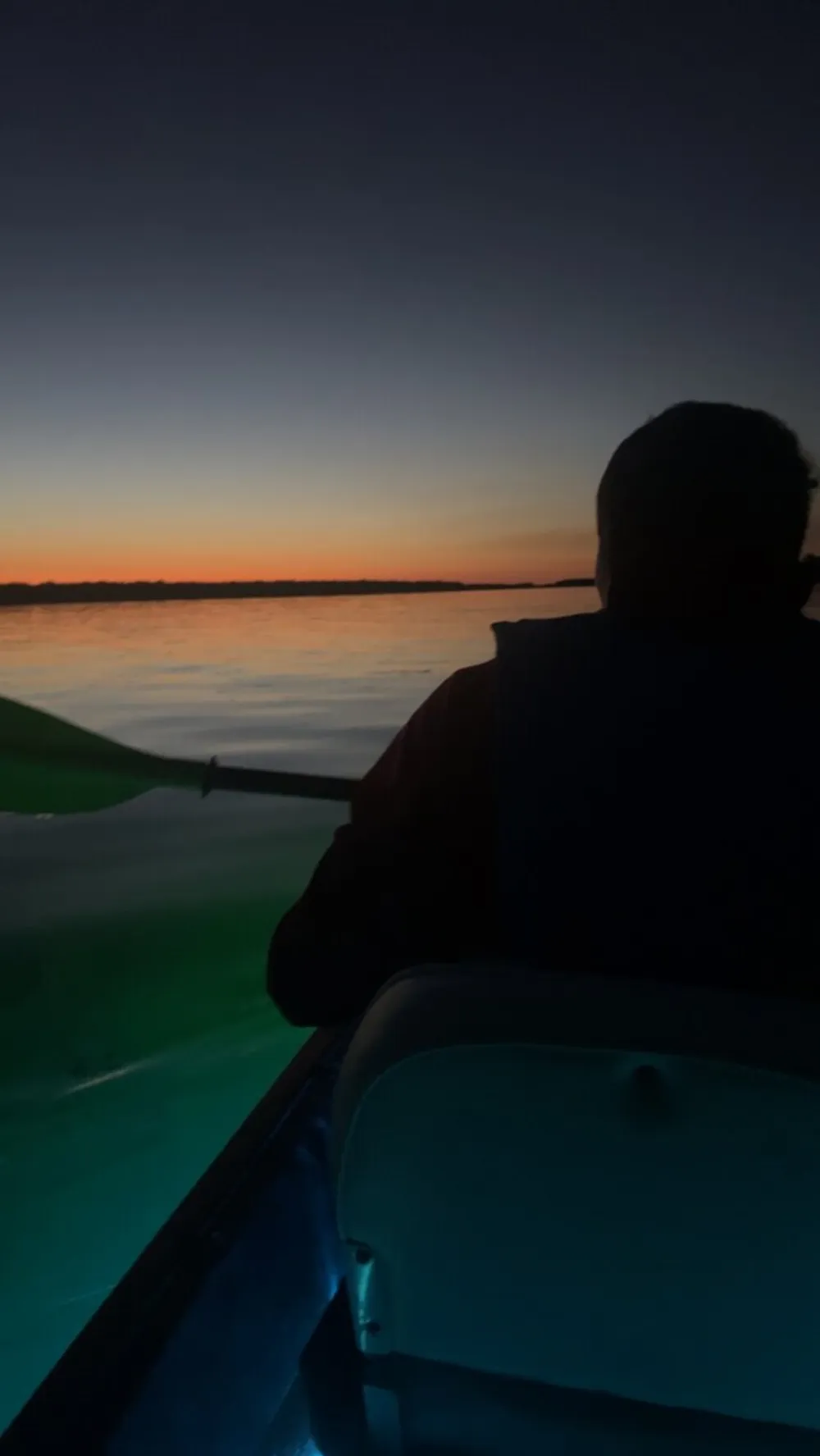 A person is seen paddling a boat at dusk silhouetted against a colorful sunset reflected on calm waters