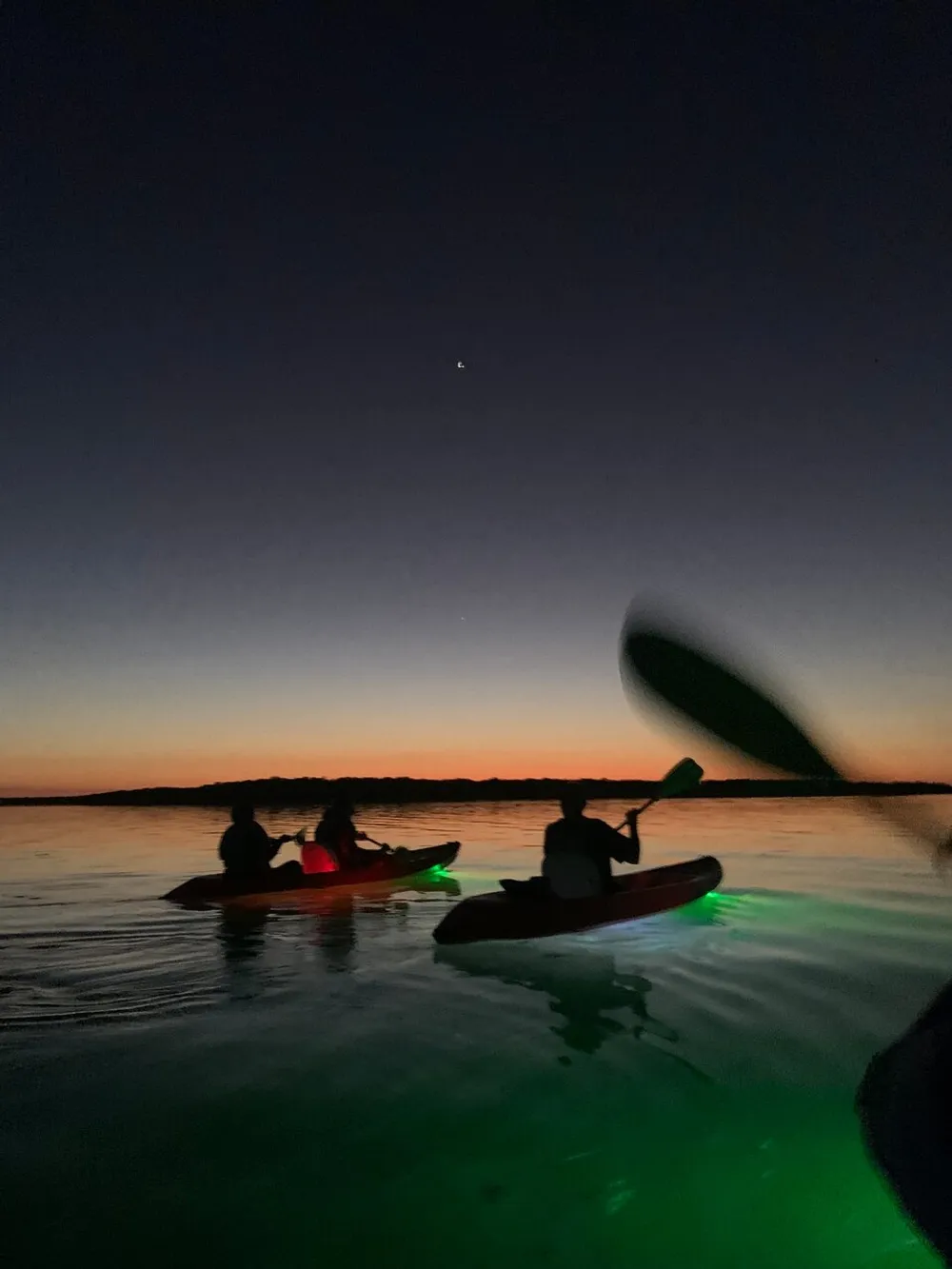 People are kayaking on calm waters against a vibrant sunset sky with a visible star above