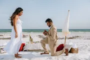 A man is kneeling on one knee on a sandy beach, appearing to propose to a woman standing in front of a romantic setup with a table, champagne, and an ocean backdrop.