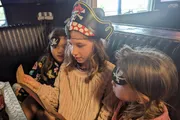 Three children, two wearing pirate hats, are looking intently at a menu in a restaurant booth.