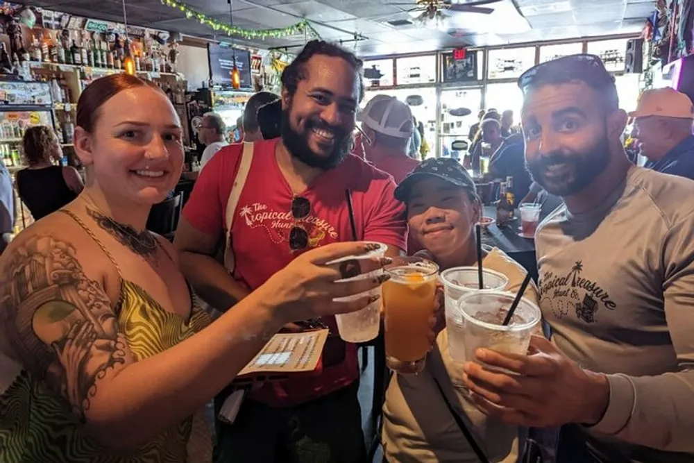 Four people are smiling and toasting with drinks in a crowded bar