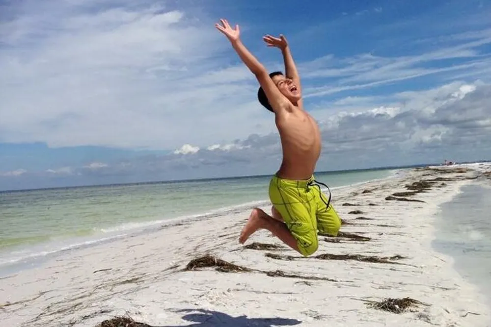 A joyful boy is jumping high on a sunny beach with clear blue skies