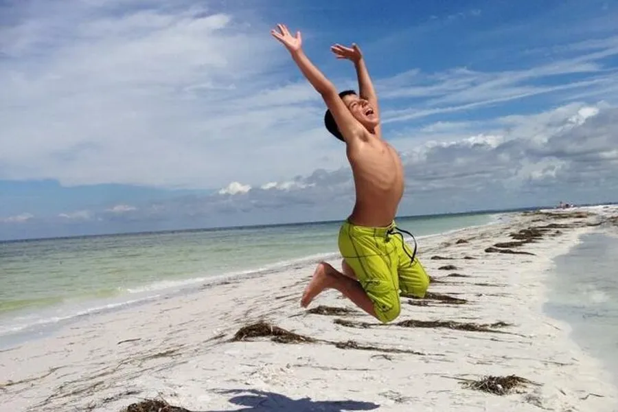 A joyful boy is jumping high on a sunny beach with clear blue skies.
