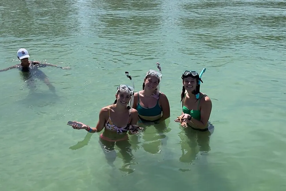 Three people are smiling at the camera while standing in clear water wearing snorkeling gear with another person swimming in the background