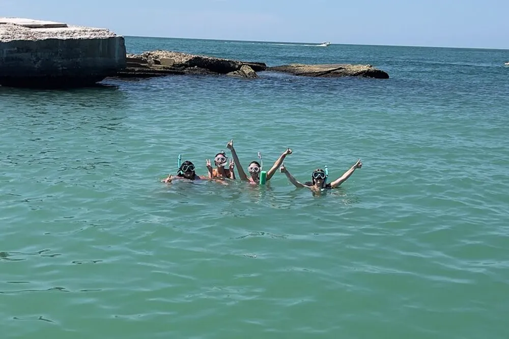 A group of four swimmers wearing snorkeling gear is waving cheerfully in clear shallow waters by the seaside