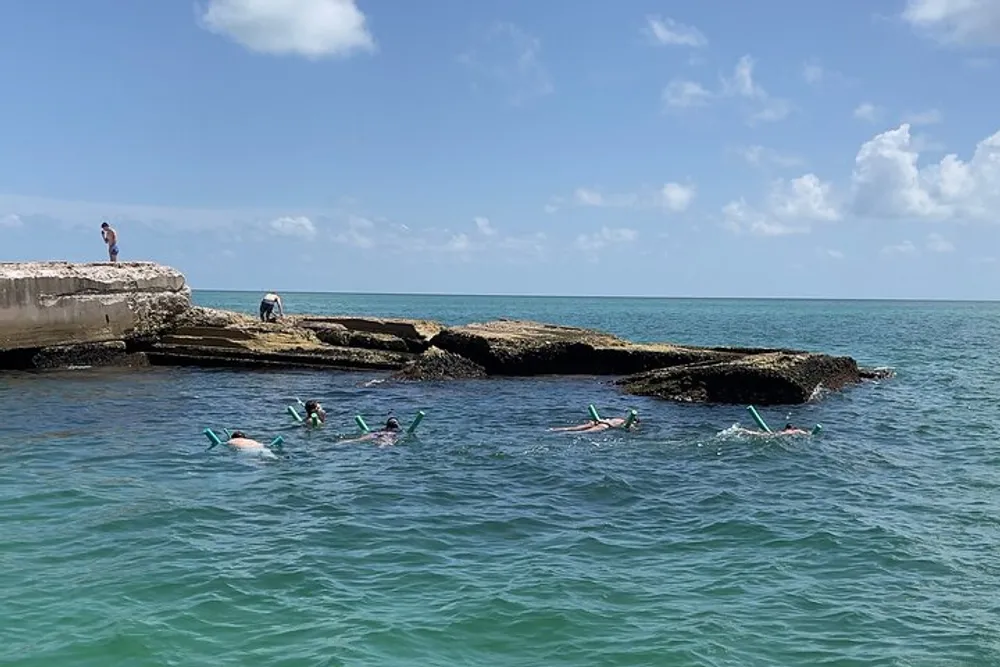 A group of snorkelers explores the waters near a rocky outcrop with a couple of people standing above on a sunny day
